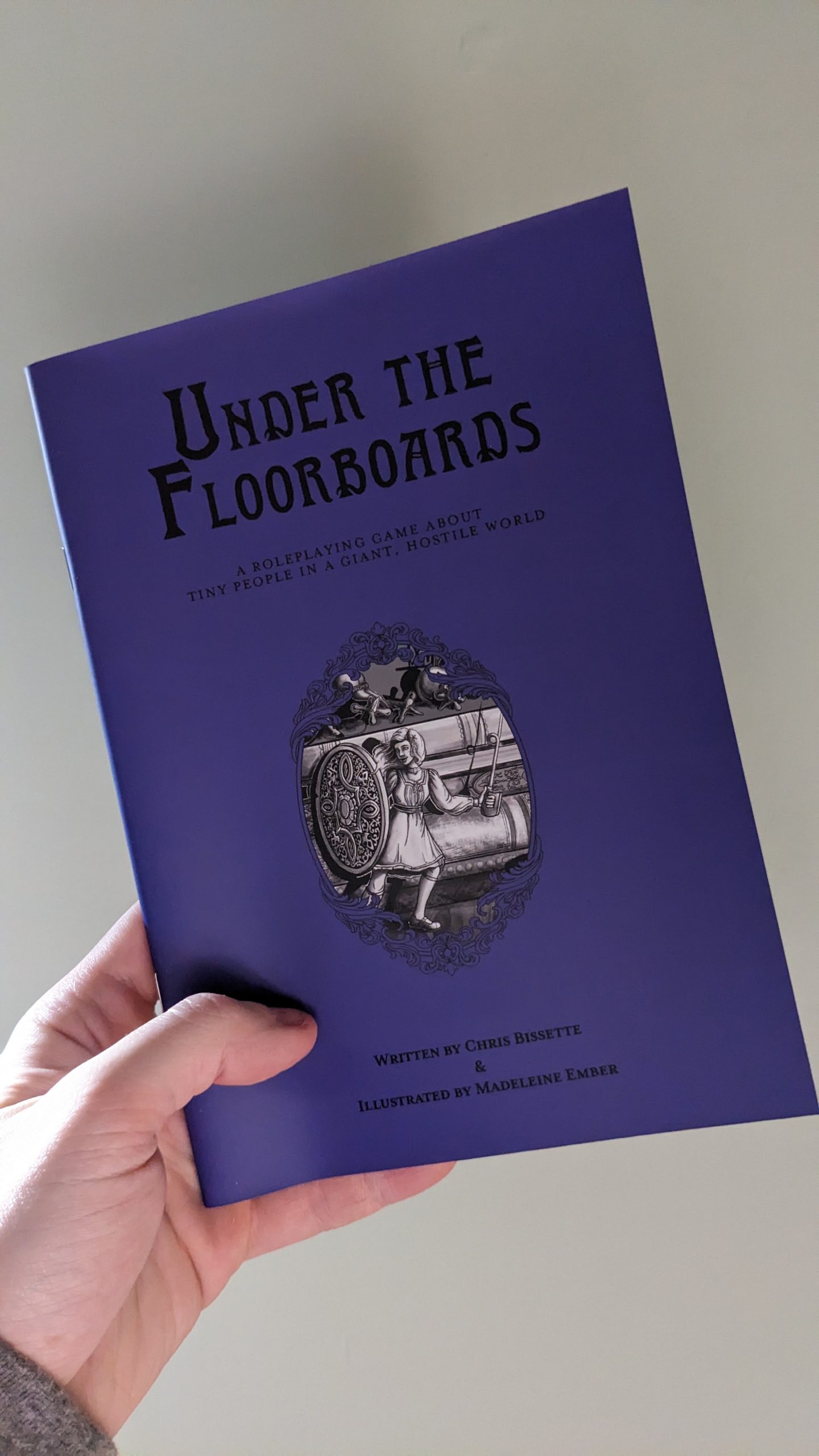 A purple A5 booklet with a black and white illustration of a tiny woman wielding a needle as a sword. The title reads "Under The Floorboards"