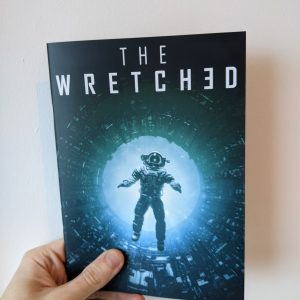 A hand holds an A5 zine against a white background. The title on the zine reads "The Wretched". On the cover, a silhouette of an astronaut falls backwards into a glowing green and blue void in the middle of a maze of industrial pipework.