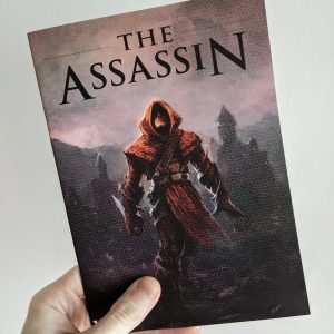 A hand holding a print zine against a white wall.. The cover shows a hooded figure in red robes holding a large knife, walking away from a city skyline. The title reads "The Assassin".