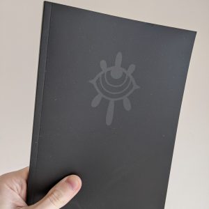 A hand holding a black softback book against a white wall. The cover is solid black, with a dark grey sigil that resembles a weeping eye.