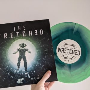A hand holds a vinyl record against a white background. The record is sticking out of its sleeve. The sleeve shows an astronaut falling backwards into a glowing white void, with the title THE WRETCHED above them. The record itself is marble with two different shades of green. The label holds a sci-fi logo that reads THE WRETCHED.