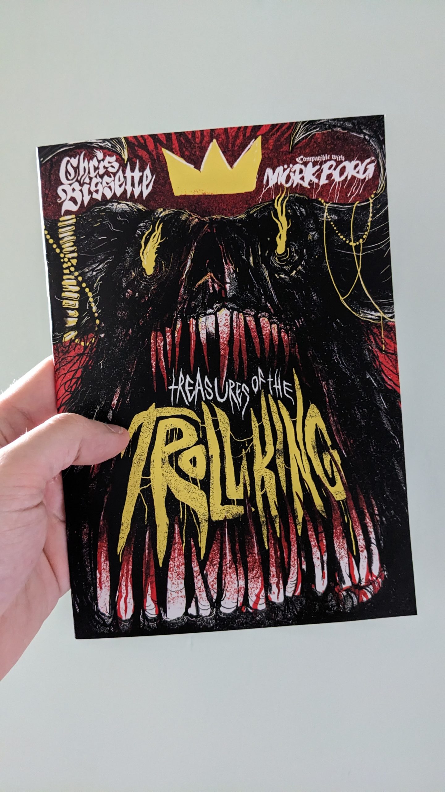 An A5 zine held against a plain white background. The cover shows a dark dreature with an abundance of teeth, dripping in blood. In the space between the teeth yellow text reads "Treasures Of The Troll King". The creature wears a yellow crown.
