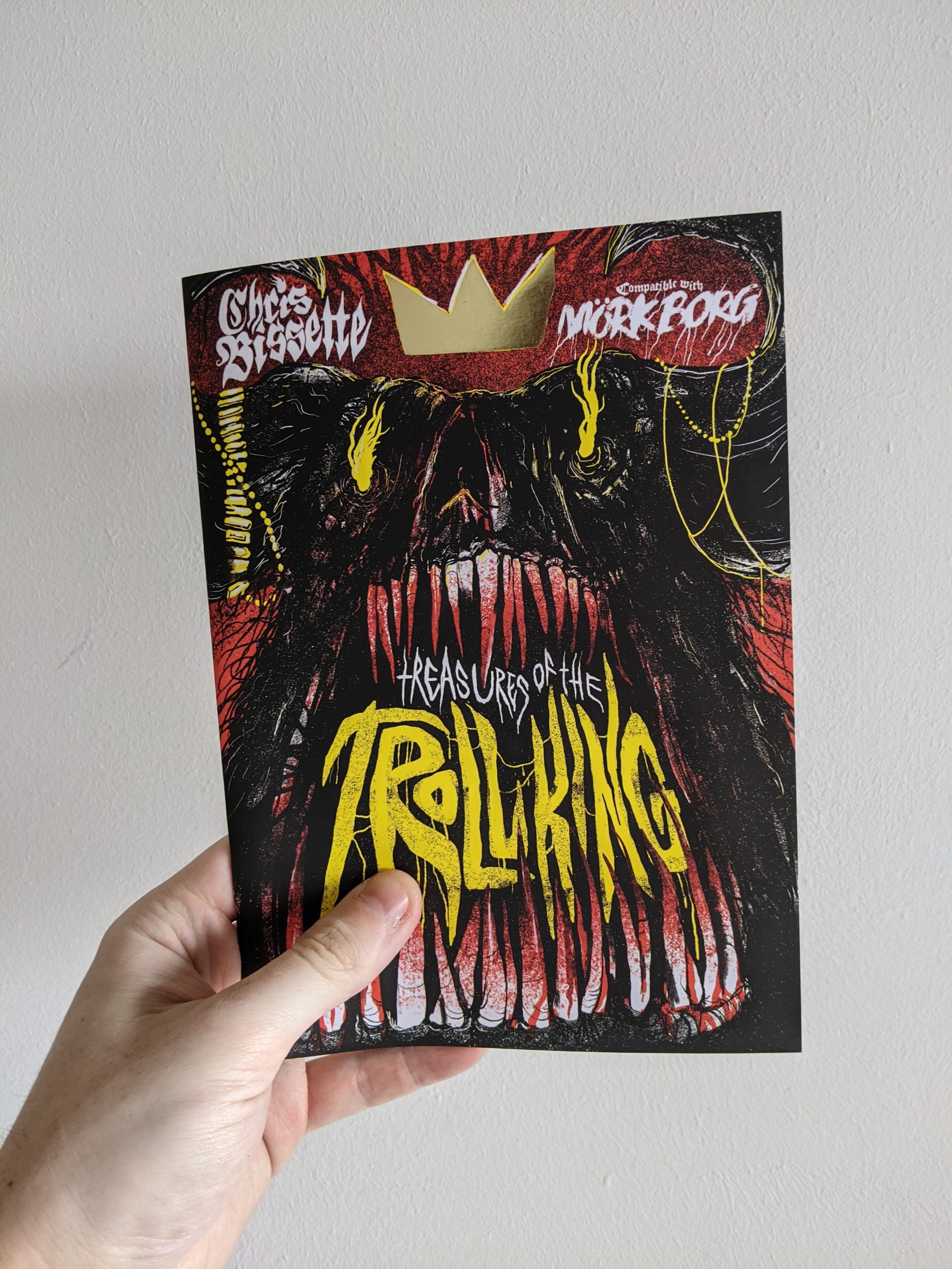 An A5 zine held against a plain white background. The cover shows a dark dreature with an abundance of teeth, dripping in blood. In the space between the teeth yellow text reads "Treasures Of The Troll King". The creature wears a crown that is printed in gold foil.