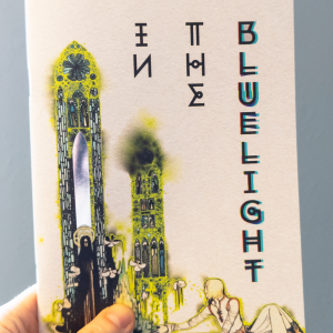 An A5 zine held in front of a plain blue wall. The cover is mostly white and shows an illustration of a reclining figure reaching bac towards a robed character standing in front of stained glass windows. One of the windows is printed with silver foil. The title reads "In The Bluelight" in an angular, almost runic font.