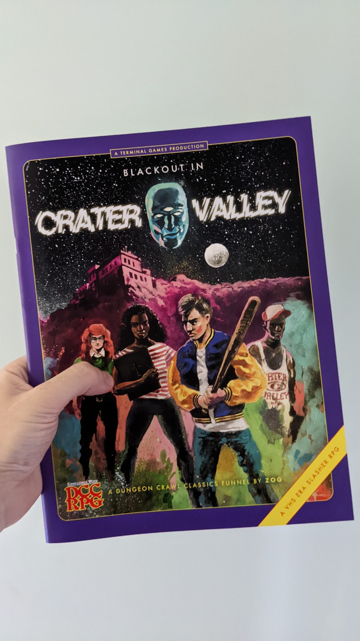 A softcover A4 book. The cover resembles an 80s VHS tape. The title, in digitally distorted font, reads "Blackout In Crater Valley" above an illustration of a group of teenagers armed with baseball bats.
