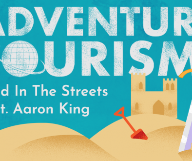 A blue banner showing golden sand dunes with a sandcastle in the background. A large sword is stabbed into the sand in the foreground. A grungey white title reads "Adventure Tourism". A clean white subtitle gives the title of the episode, "Wild In The Streets feat. Aaron King"