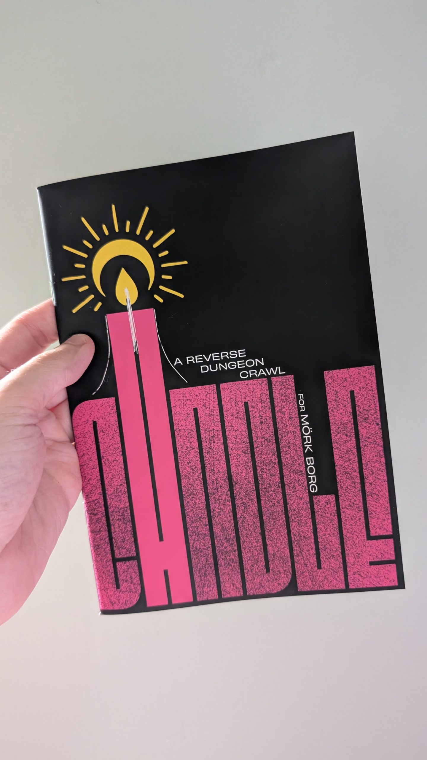 An A5 zine held in front of a plain background. The cover is text-based, with large pink type that reads CANDLE. The 'A' is elongated and has a flame above it.